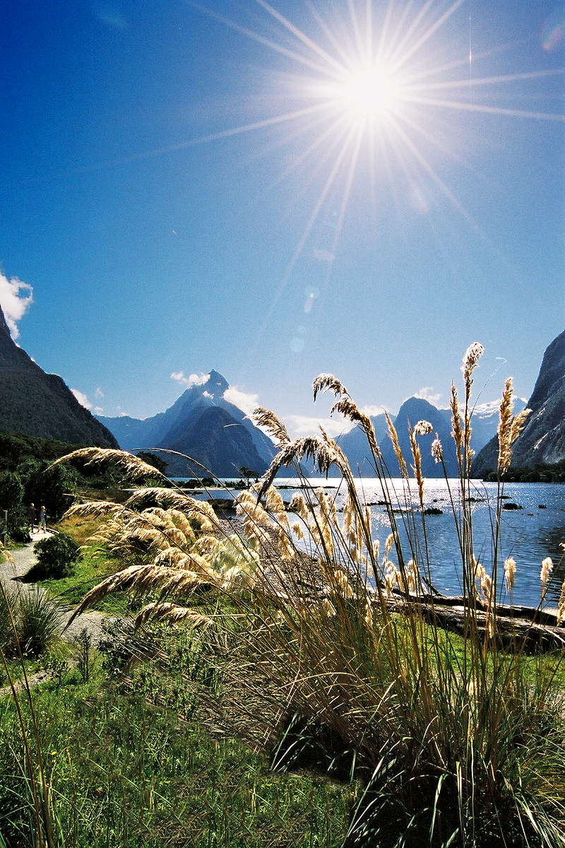 Fiordland — Milford Sound: Mitre Peak with sunburst and walkers.